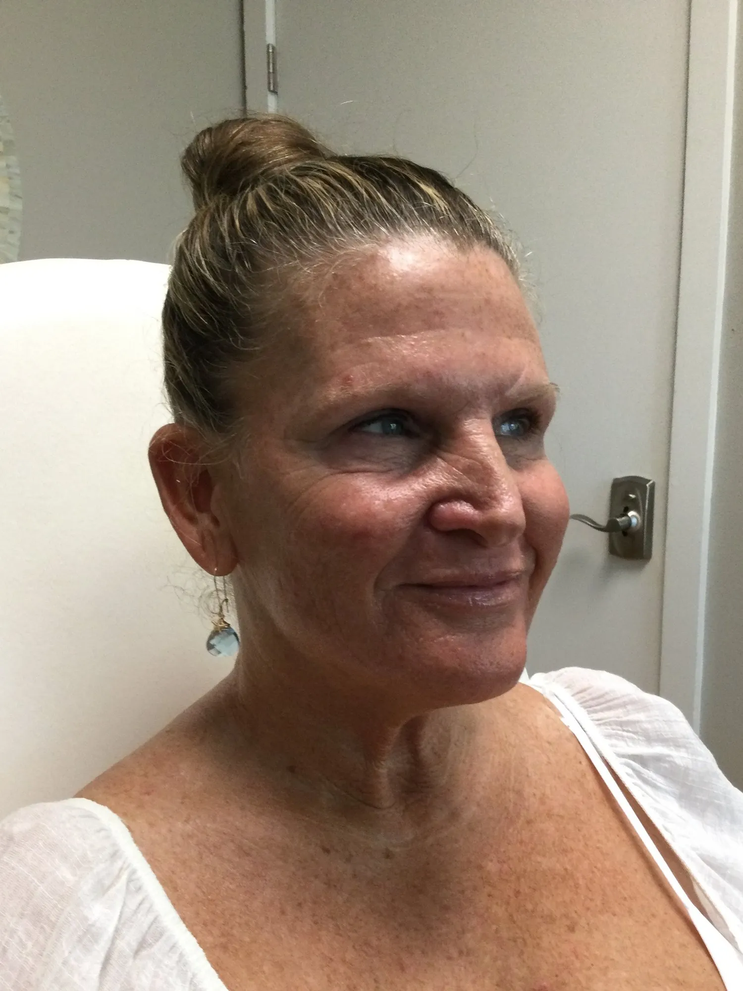 Smiling woman with hair in a bun wearing a white top and a dangling earring, sitting indoors near a closed door.