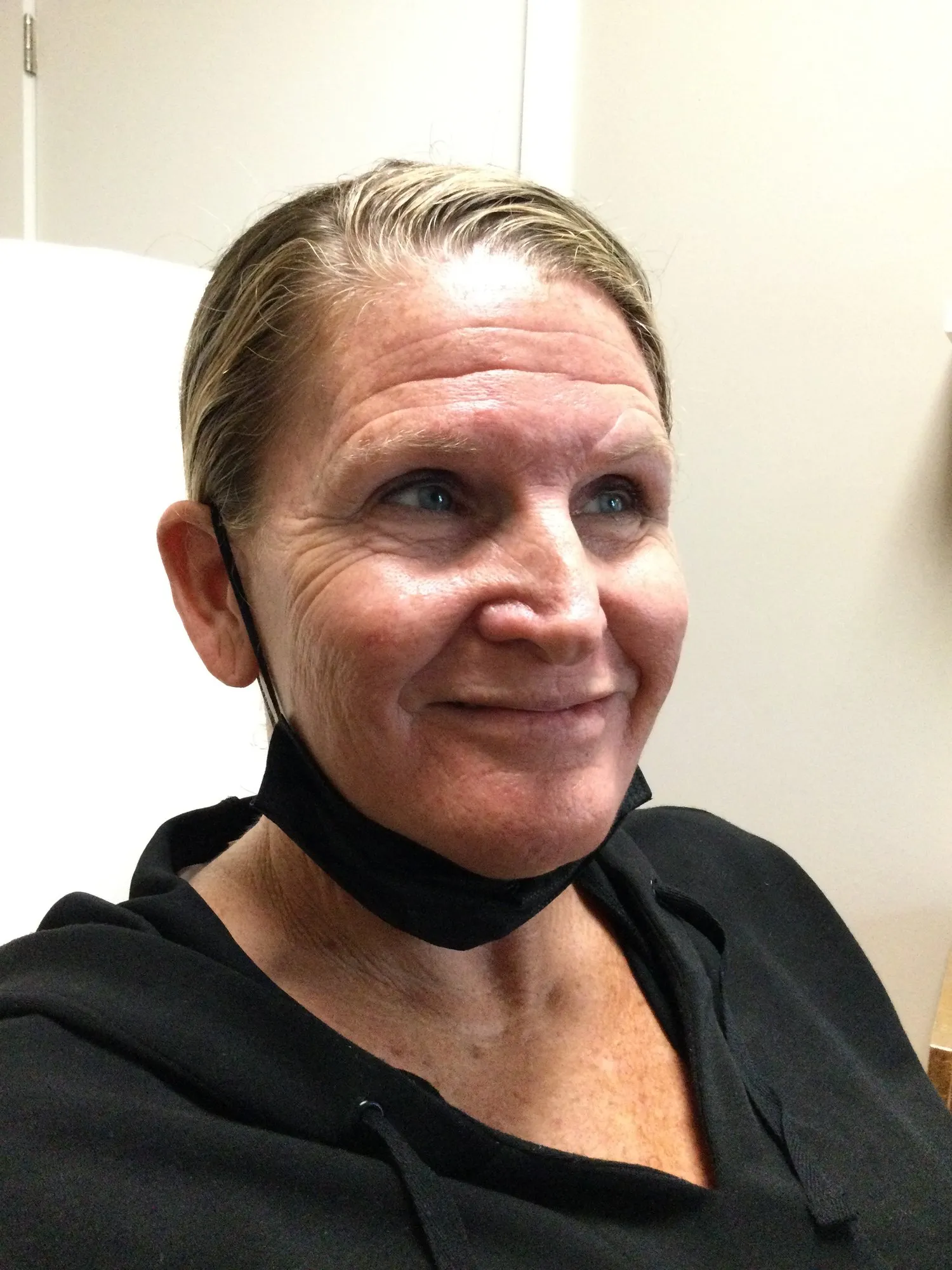 Smiling woman with light hair pulled back, wearing a black hoodie and a black face mask pulled below her chin, sitting indoors.