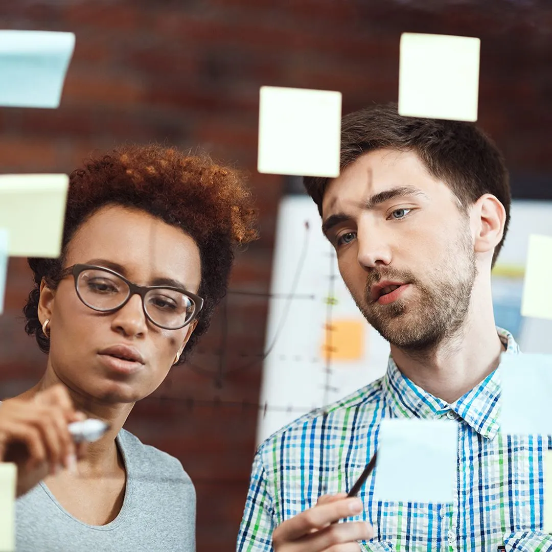 Two colleagues looking thoughtfully at sticky notes on a wall, one wearing glasses and a gray top and the other in a plaid button-down shirt, with a brick wall background.