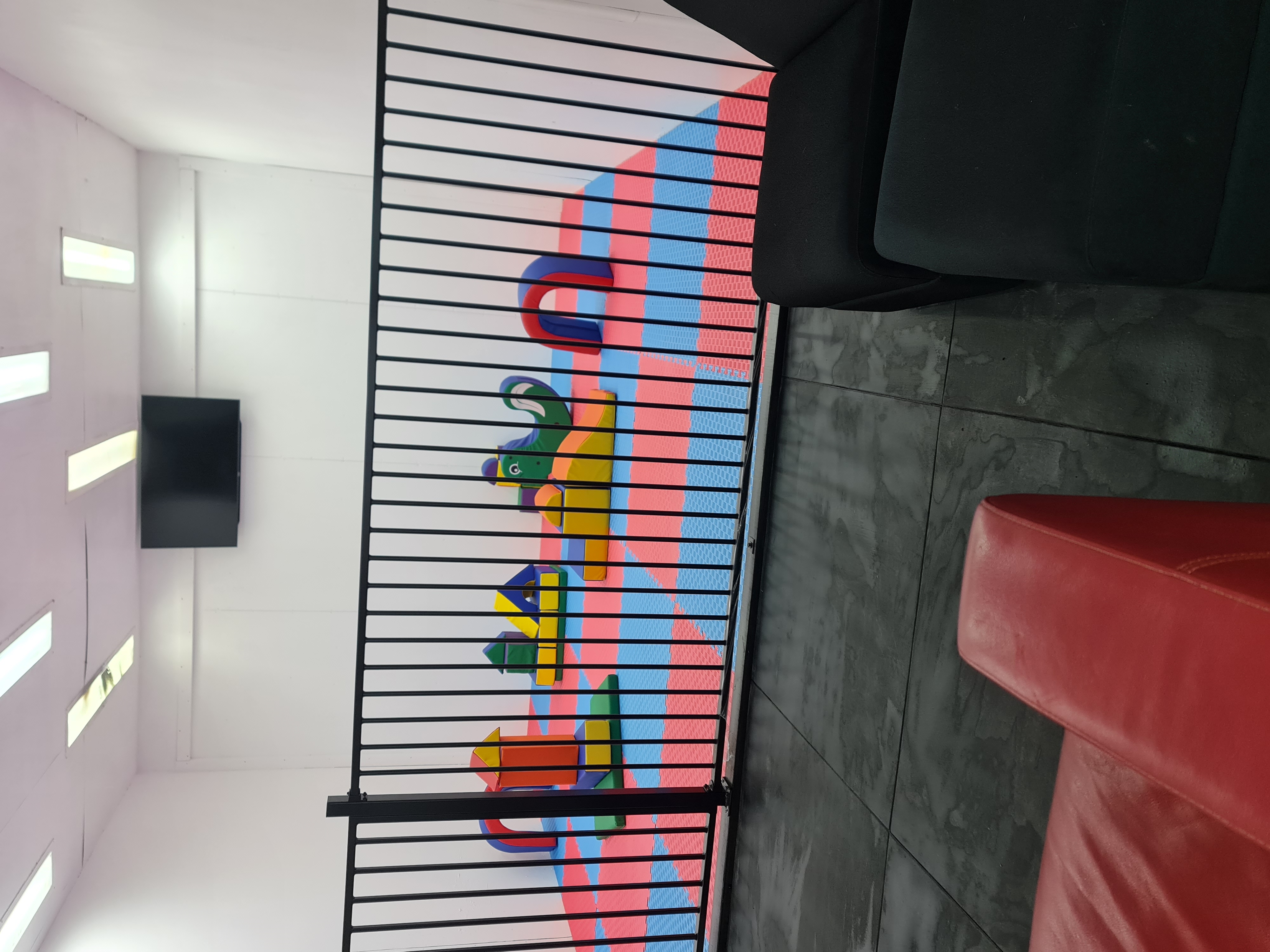 The finished MFF playroom: soft play blocks and colourful mats behind the child-safety fence, with a movie screen and sofas alongside