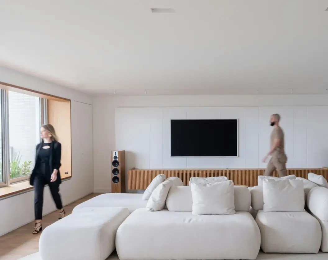 Modern living room with white sectional sofa, wooden TV console, flat-screen TV on the wall, and two people walking past.