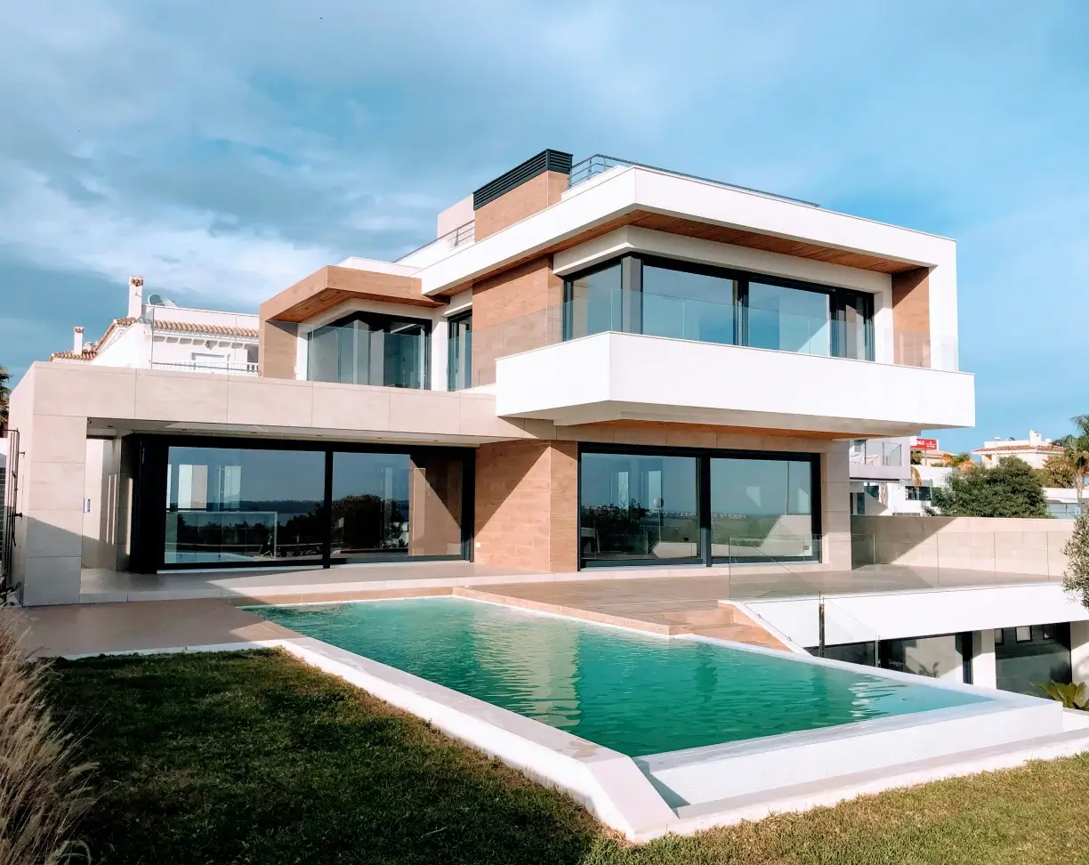 Modern two-story house with large glass windows, beige and white exterior, and a rectangular pool in front.