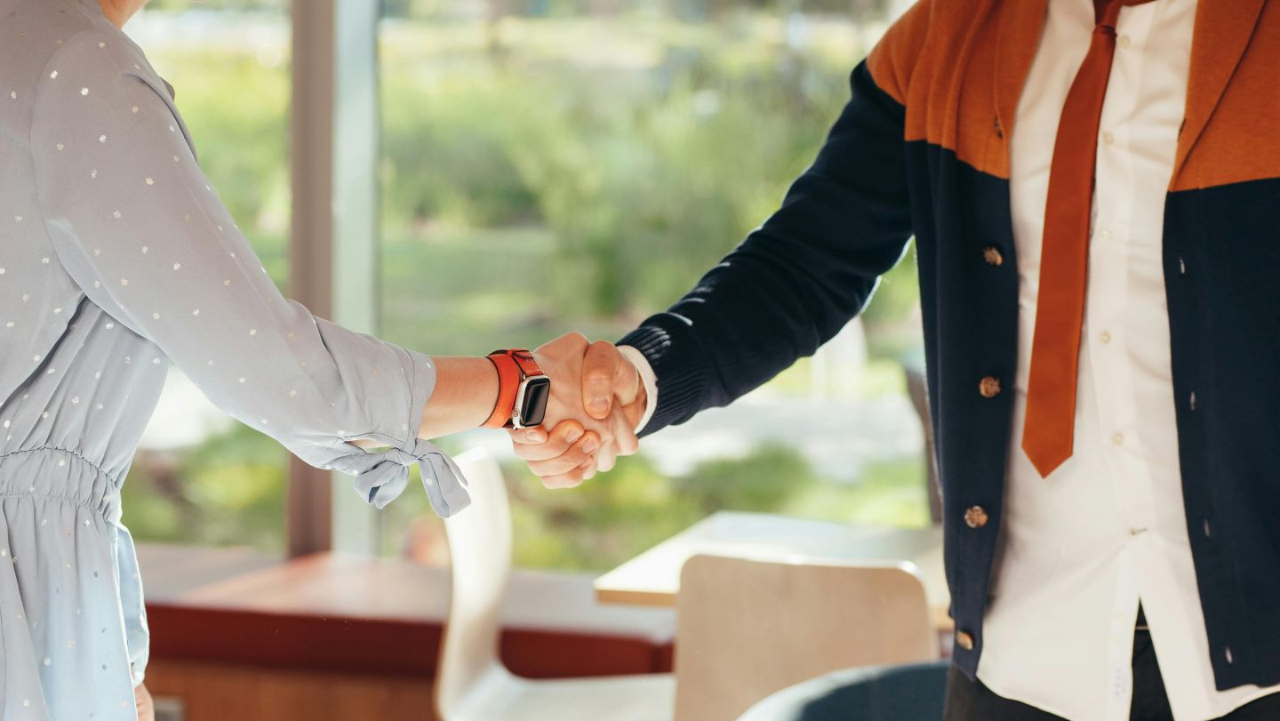 Two people shaking hands indoors near a window, one wearing a light blue dress and smartwatch, the other in a white shirt, orange tie, and navy cardigan.