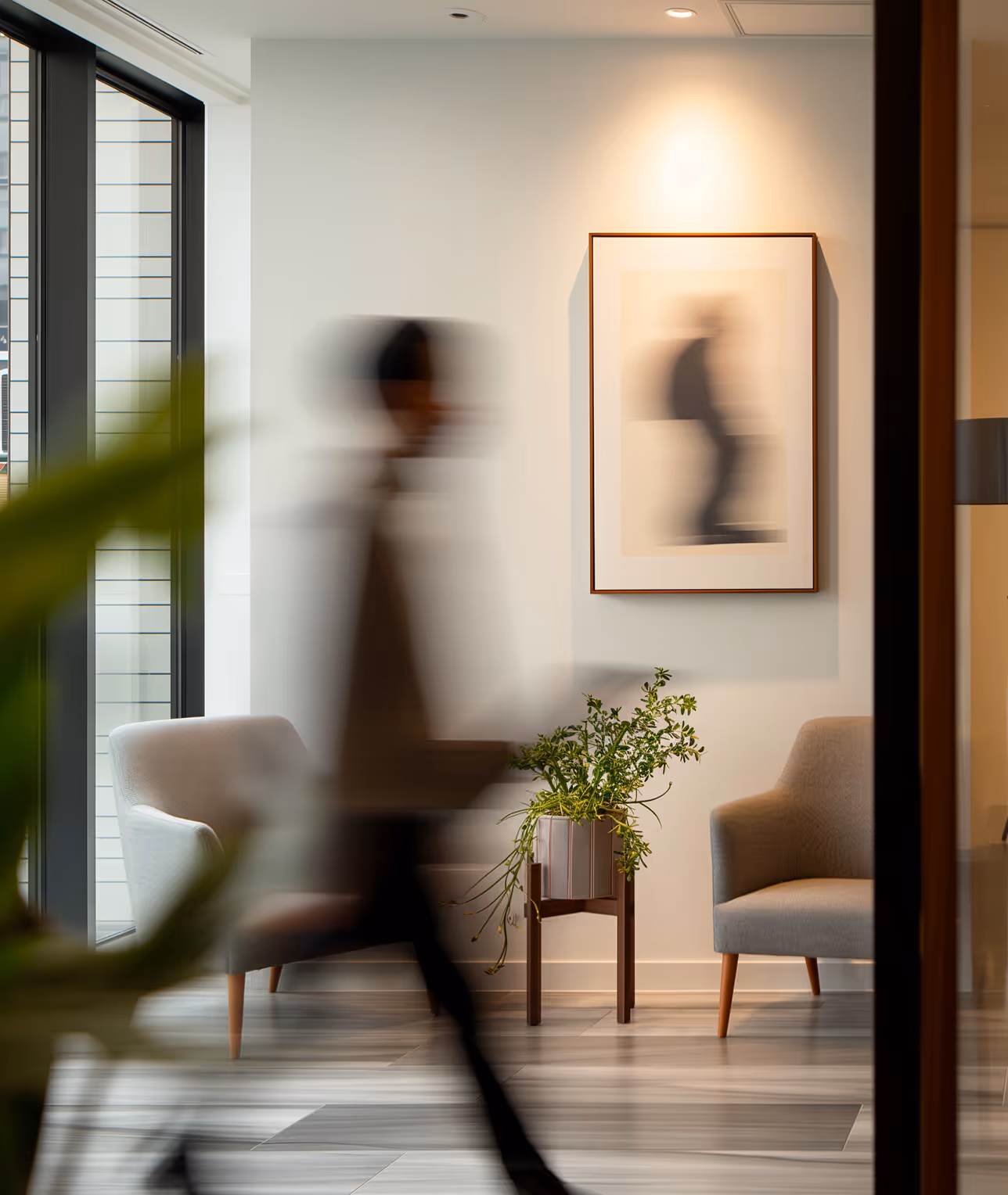 Blurred figure walking past two armchairs and a plant stand beneath a framed blurred artwork in a modern office lobby.
