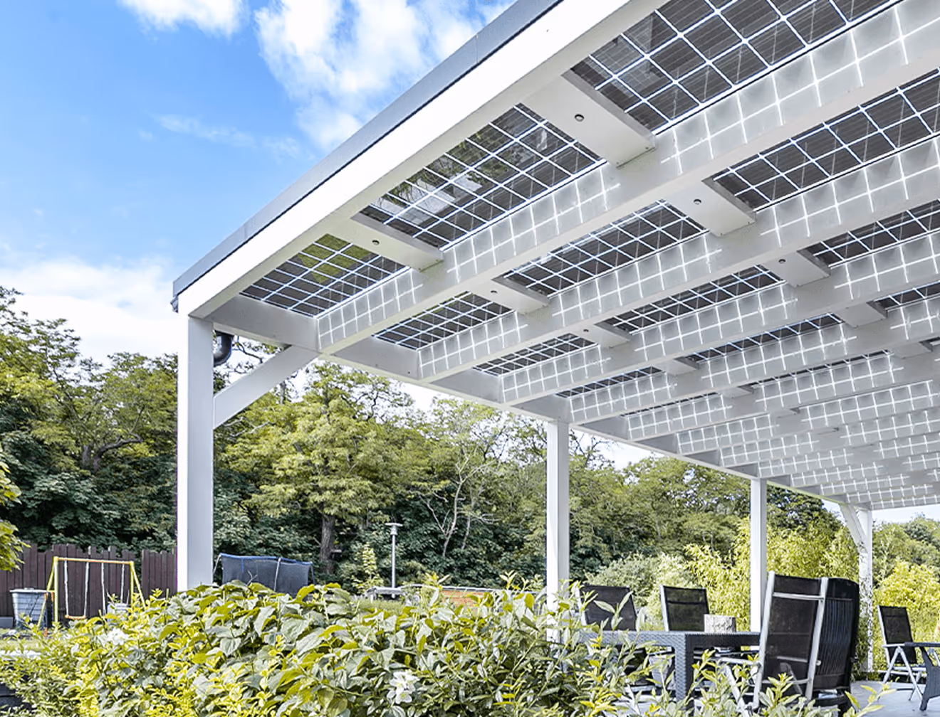 Solarüberdachung mit transparenten Paneelen über Gartenmöbeln, von Bäumen umgeben, bei klarem Himmel.