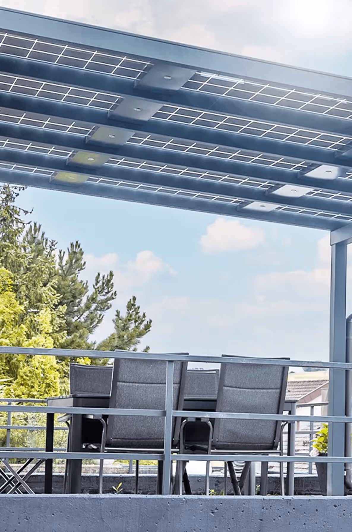 Terrasse mit modernem Solardach, Tisch und vier Stühlen, umgeben von grünen Bäumen unter blauem Himmel mit Wolken.