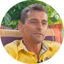 Mann mit gelbem Hemd auf bunter Couch, Hintergrund mit grünen Blättern.