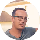 Mann mit Brille und kurzem Haar im Grünen sitzt vor einem Gebäude.