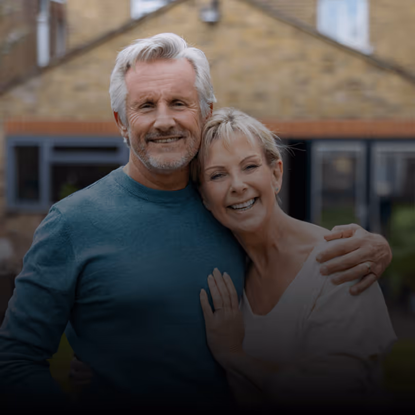 Älteres Paar lächelt im Garten vor ihrem Haus, Mann in blauem Pullover, Frau umarmt ihn, entspannte Atmosphäre.