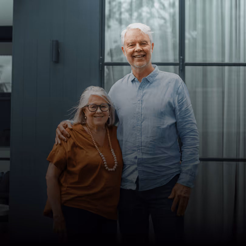 Älteres Paar lächelt, steht vor modernem Gebäude mit Glasfenster, er trägt blaues Hemd, sie orange Bluse.