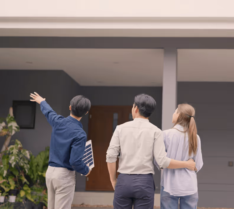 Drei Personen betrachten ein modernes Haus; ein Mann zeigt auf den Eingang, daneben ein Paar mit umarmten Händen.