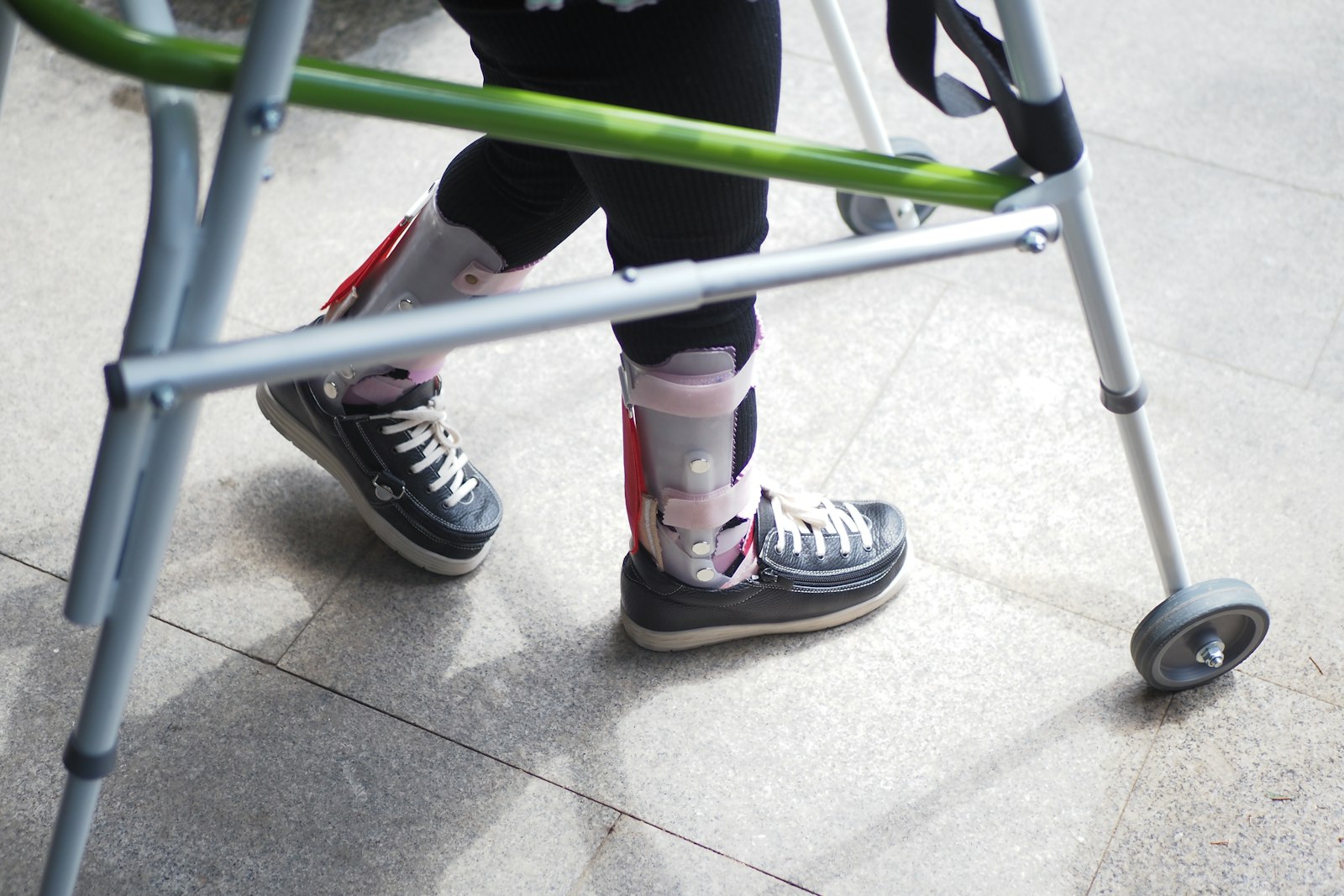 Close-up of a person's legs wearing ankle-foot orthotics and sneakers, walking with a green walker.