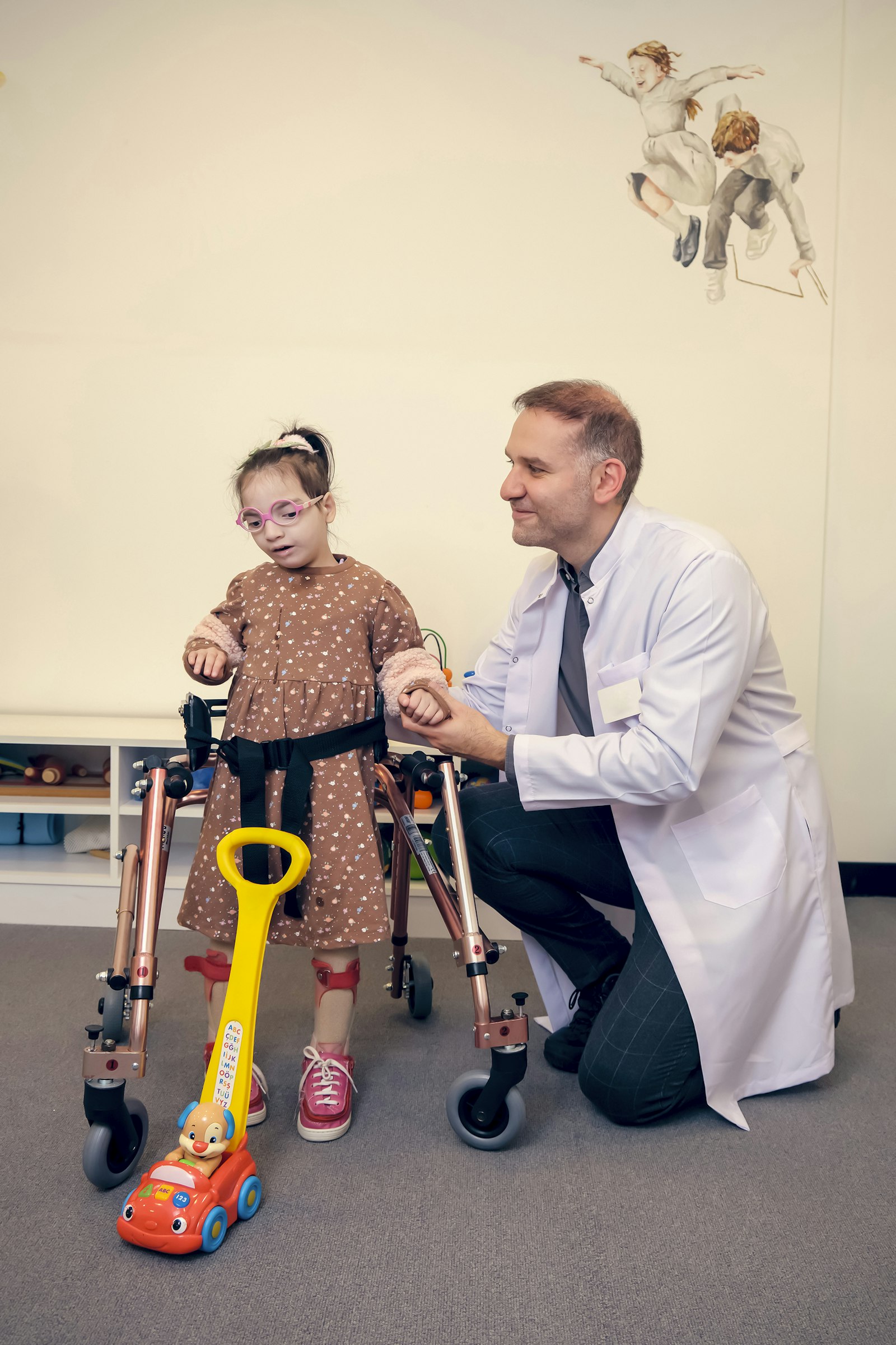 A doctor kneels beside a young girl standing in a gait trainer in a clinic room.