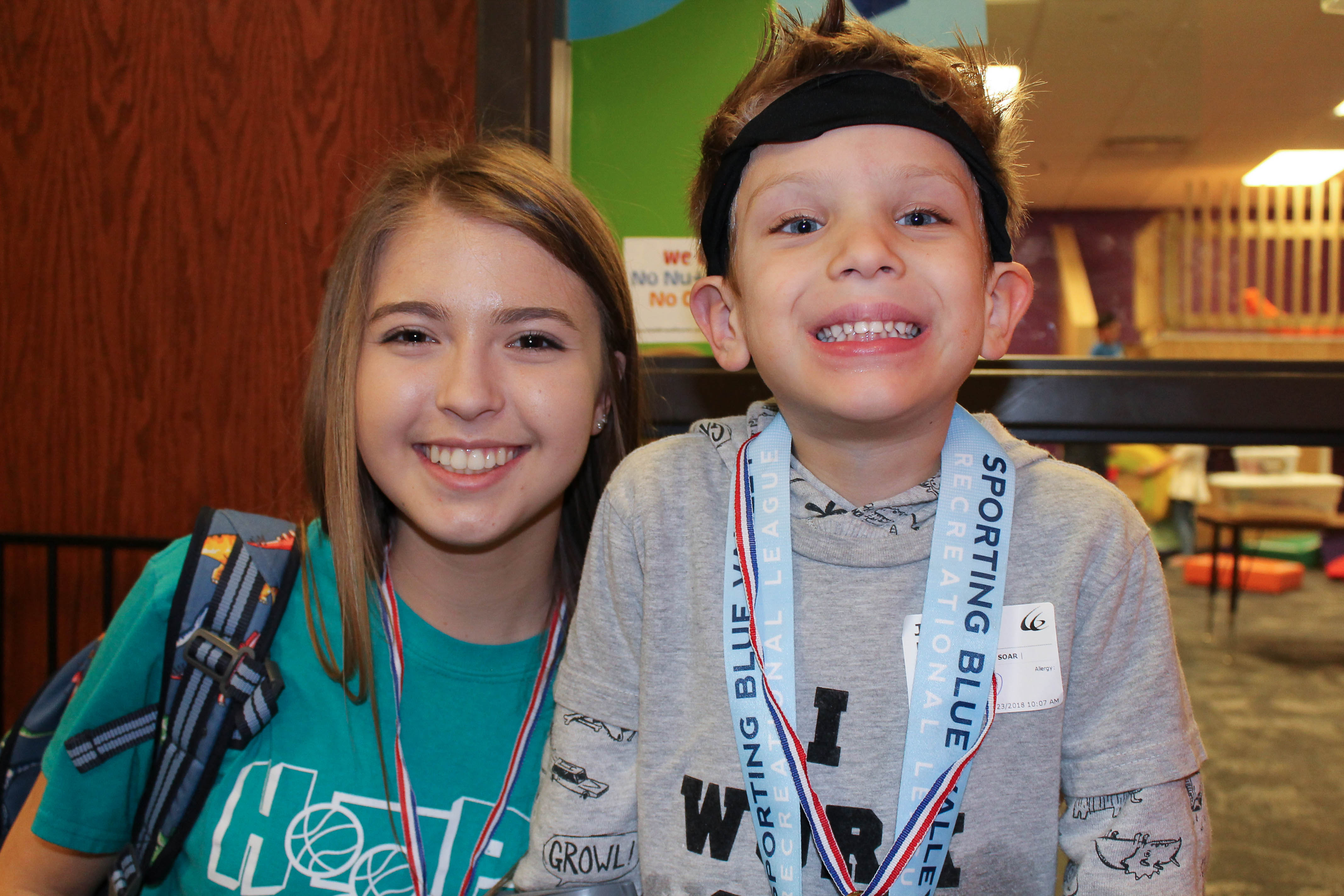 Two smiling kids pose together, one wearing a medal on a lanyard.