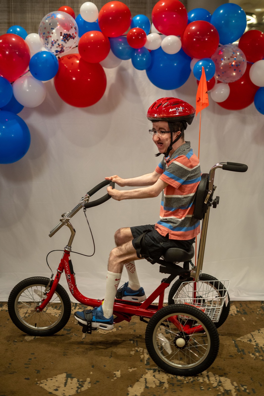 A smiling boy in a red helmet rides a red Amtryke adaptive tricycle with a back support.