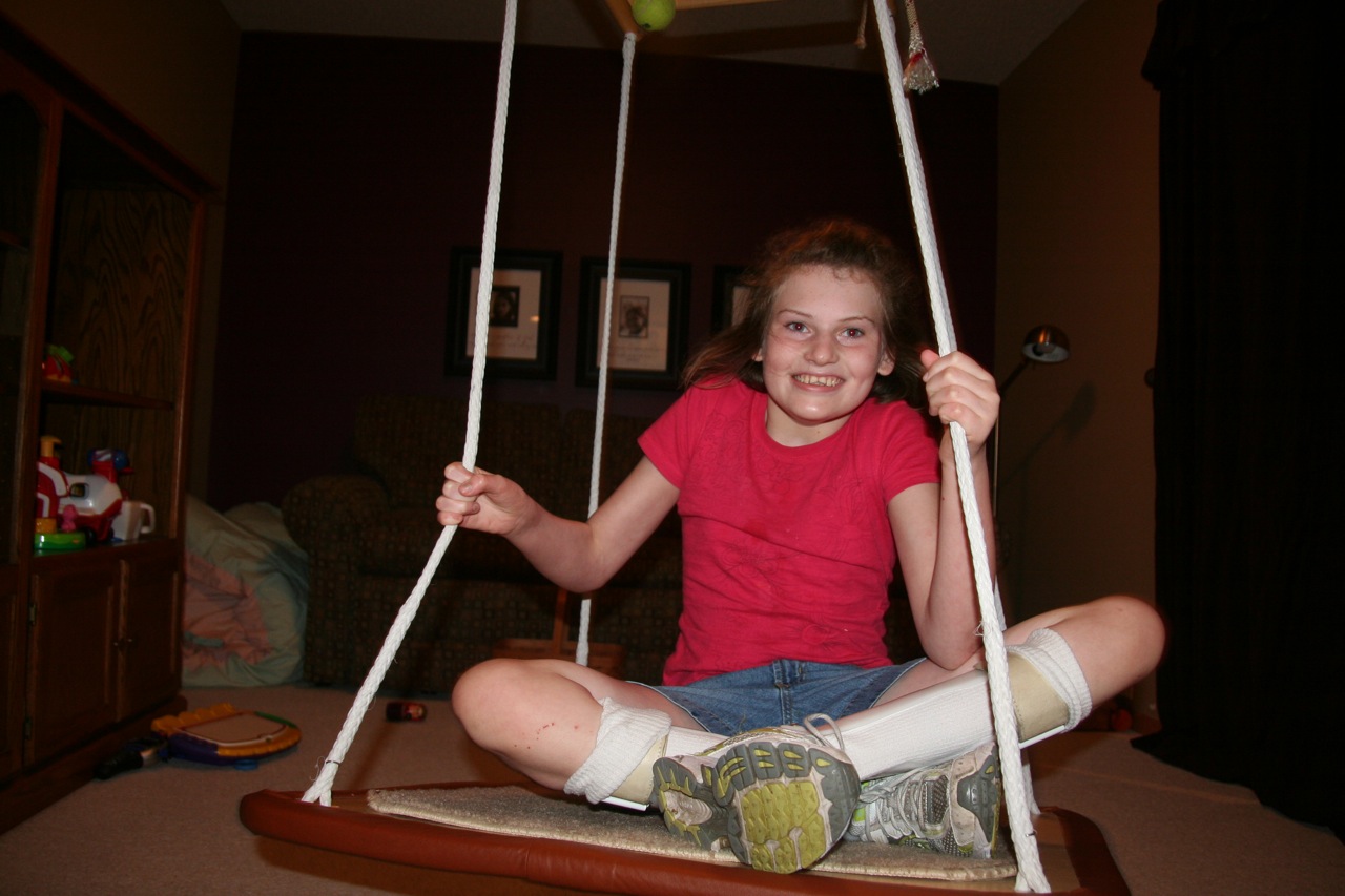 A smiling girl wearing leg braces sits on a platform swing.