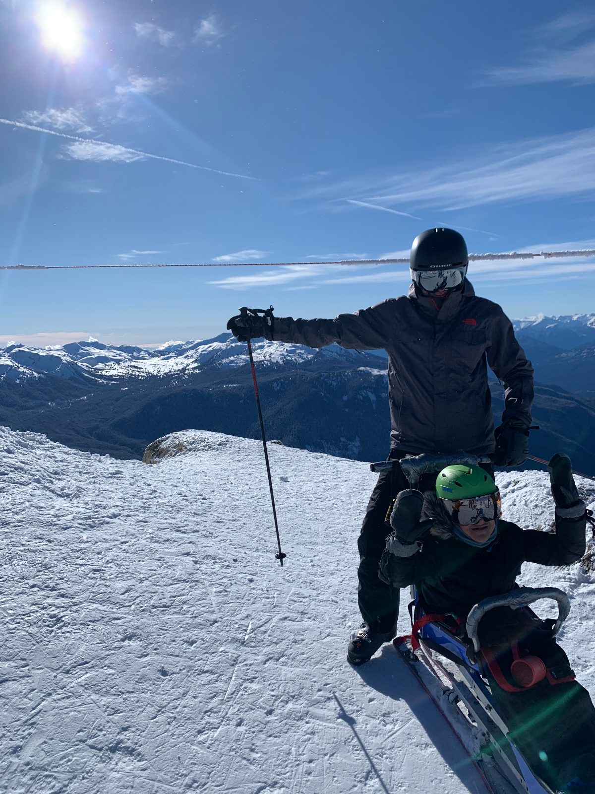 Our son in his sit-ski on a snowy mountaintop, with his dad standing behind him raising a ski pole