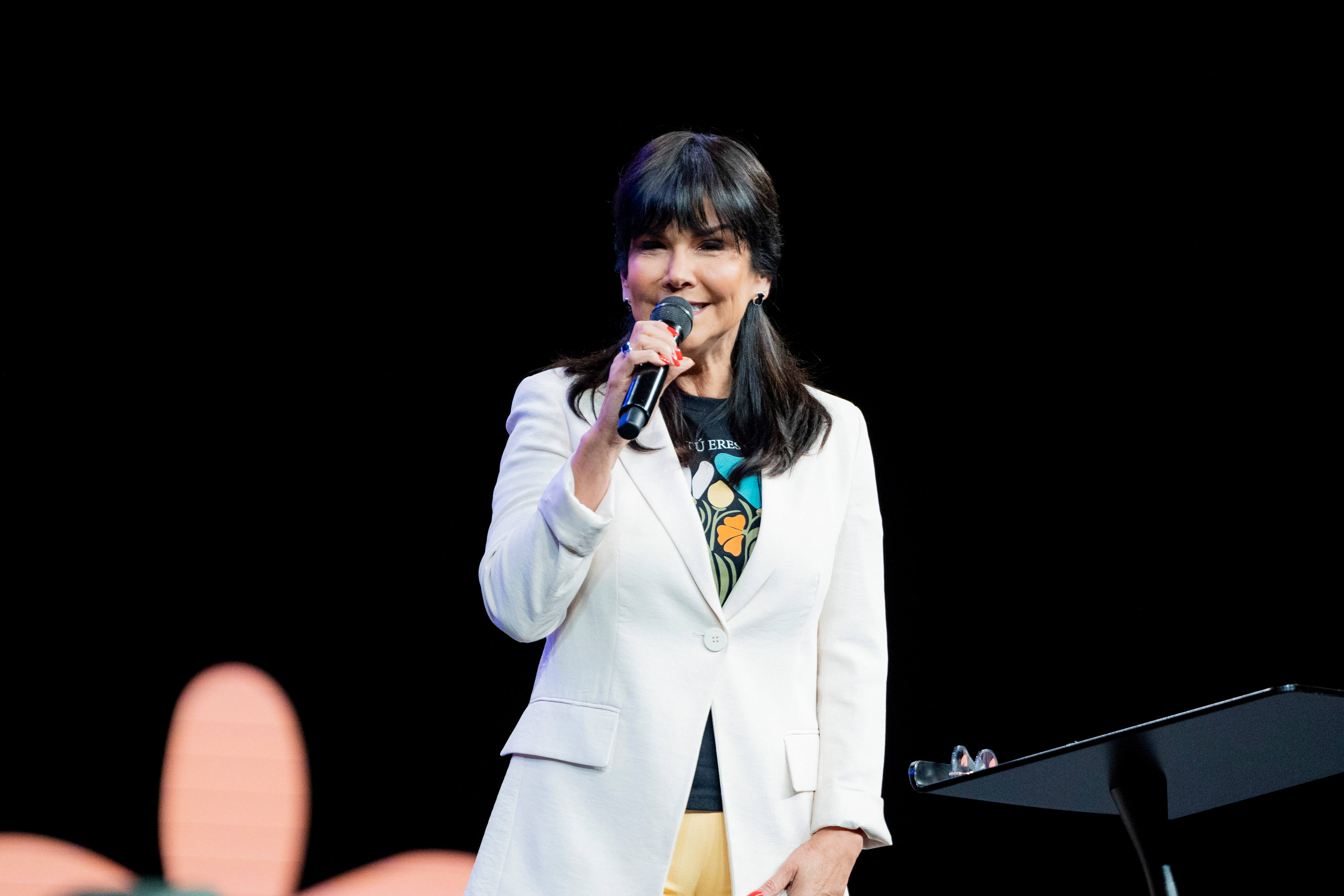 Woman with dark hair wearing a white blazer and yellow pants speaking into a microphone on stage.