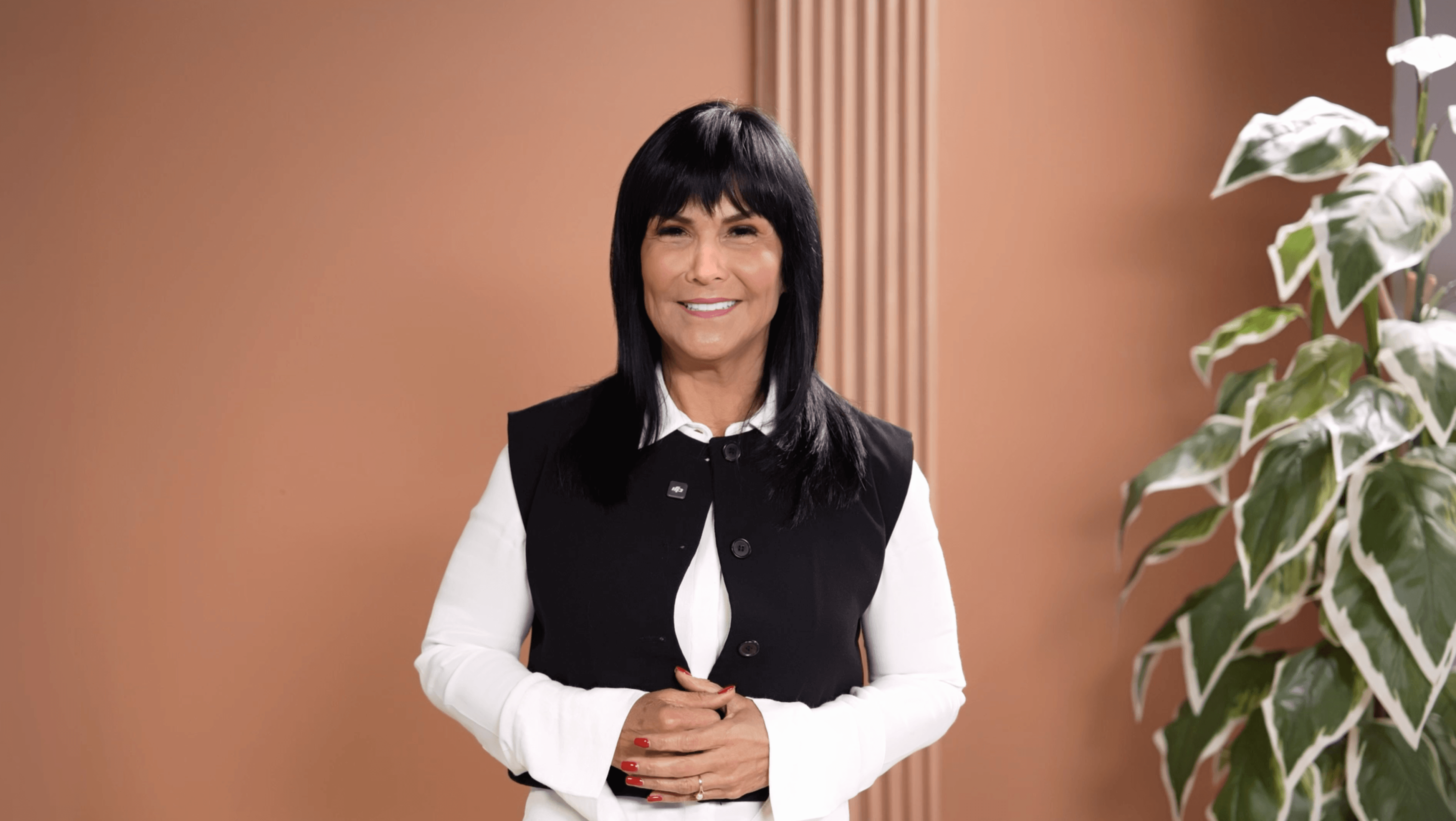 Smiling woman with straight black hair wearing a white shirt and black vest standing indoors near a plant against a beige wall.