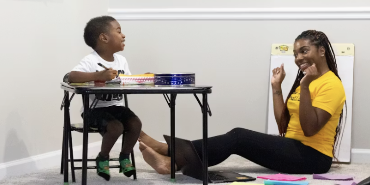 Child sitting at a small table writing with a pen while a woman sitting on the floor smiles and gestures encouragingly.