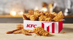 KFC box with fried chicken pieces, fries, and a dipping sauce cup on a wooden surface.