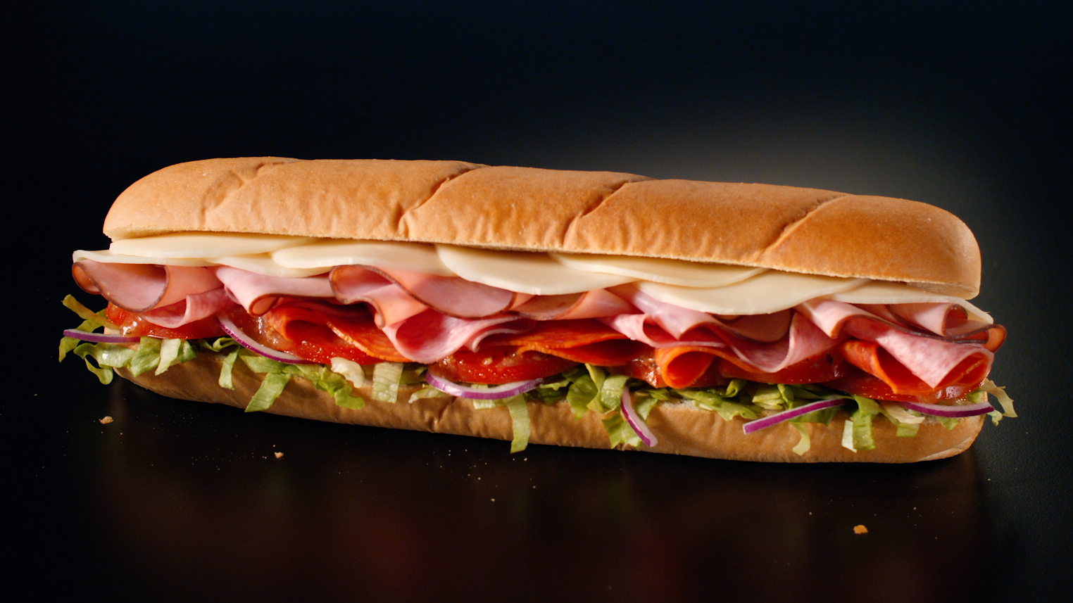 Sub sandwich with ham, pepperoni, sliced cheese, lettuce, tomato, and red onion on black background.