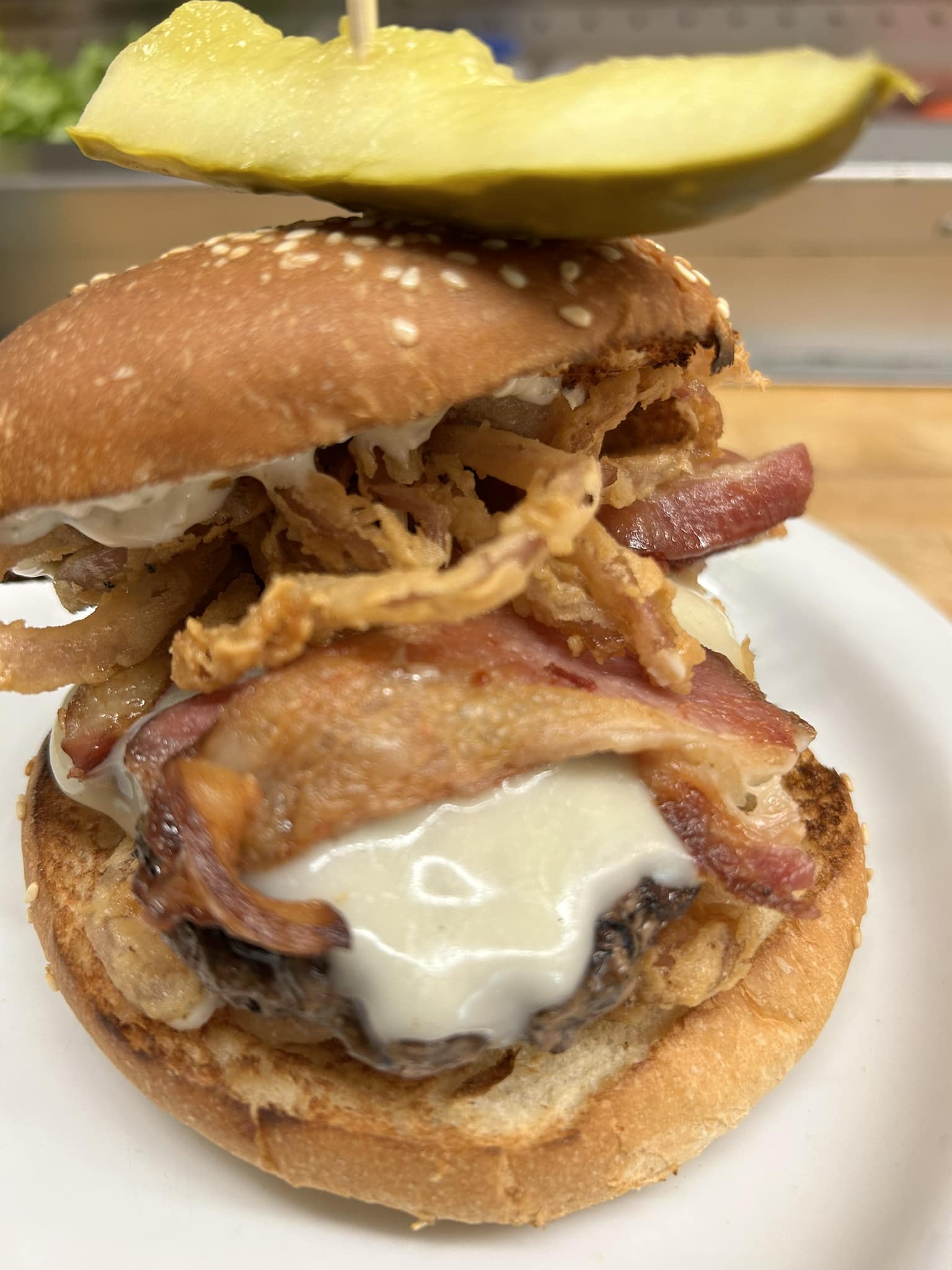 Close-up of cheese burger with bacon, crispy onion strings, and a pickle slice on a sesame bun.