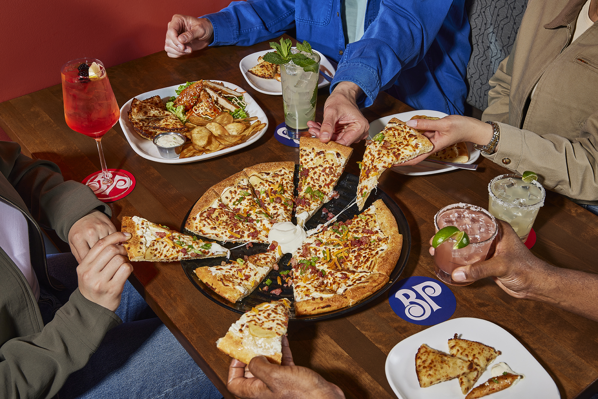 Four people sharing cheese and bacon pizza slices around a table with drinks and food.