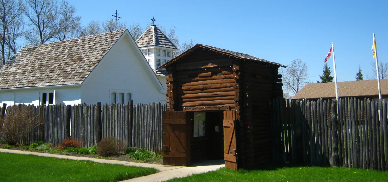 Wooden fort entrance with open double doors, white chapel, and tall wooden fence under clear sky.