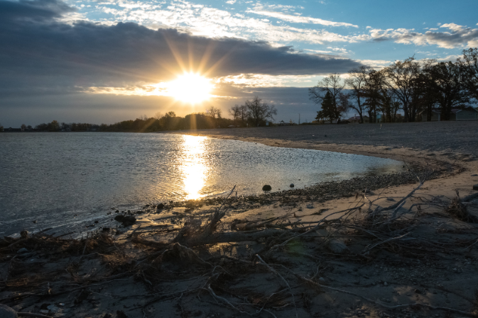 Sun setting over a calm lake with sandy shore and scattered branches in foreground.