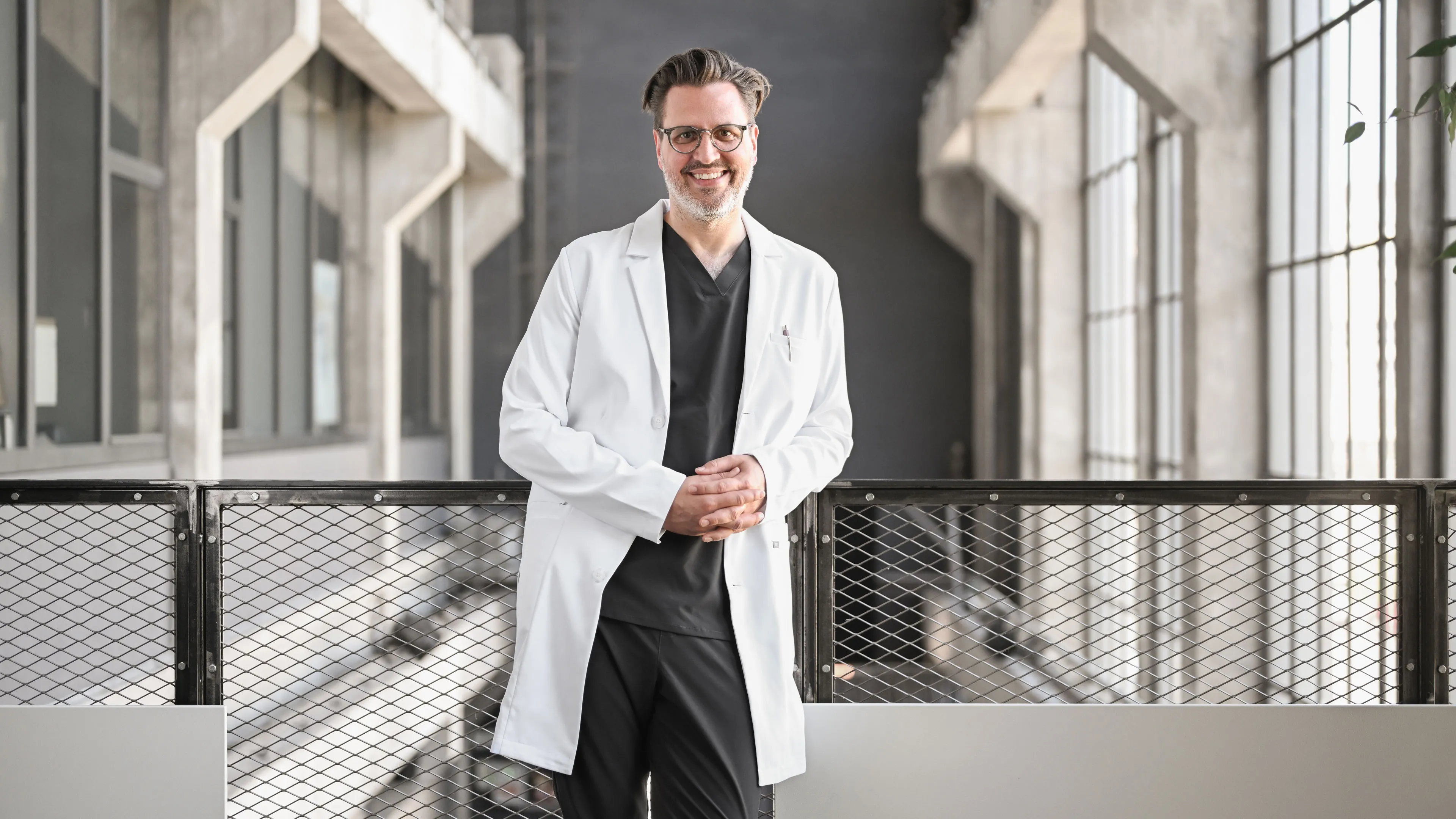 Lächelnder Arzt mit weißem Kittel und Brille steht vor modernem Geländer in hellem Flur.
