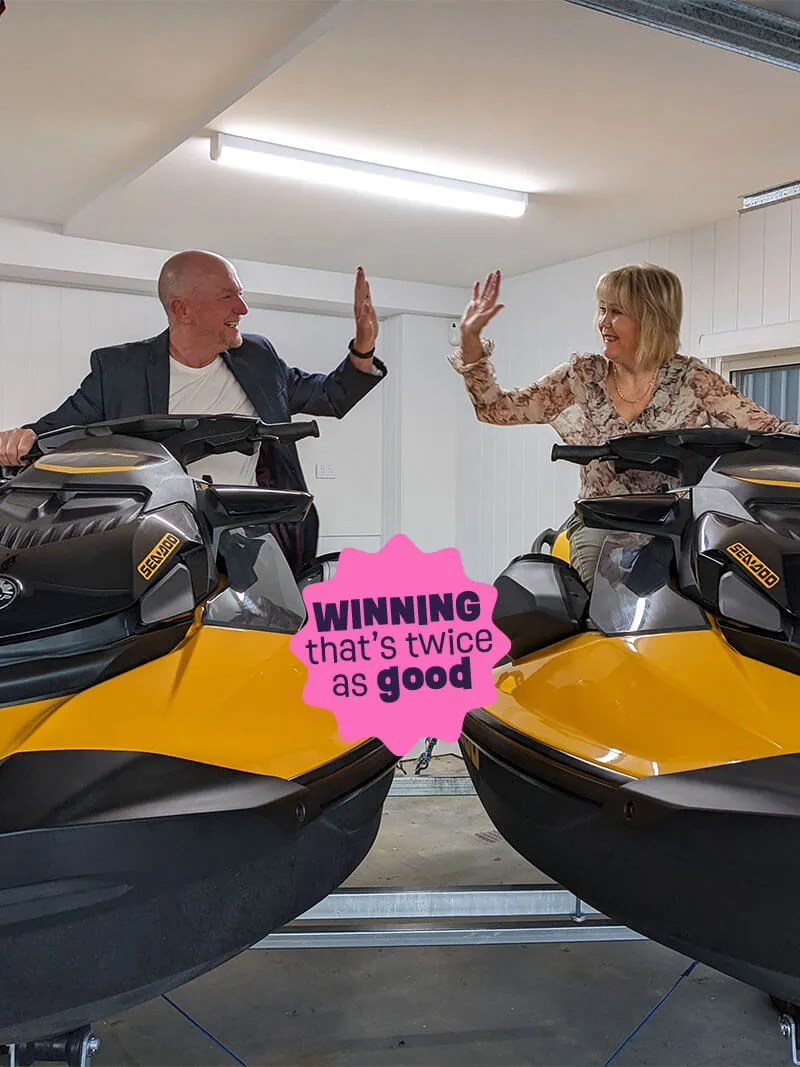A man and woman smiling and high-fiving each other while sitting on yellow Sea-Doo jet skis in a garage.