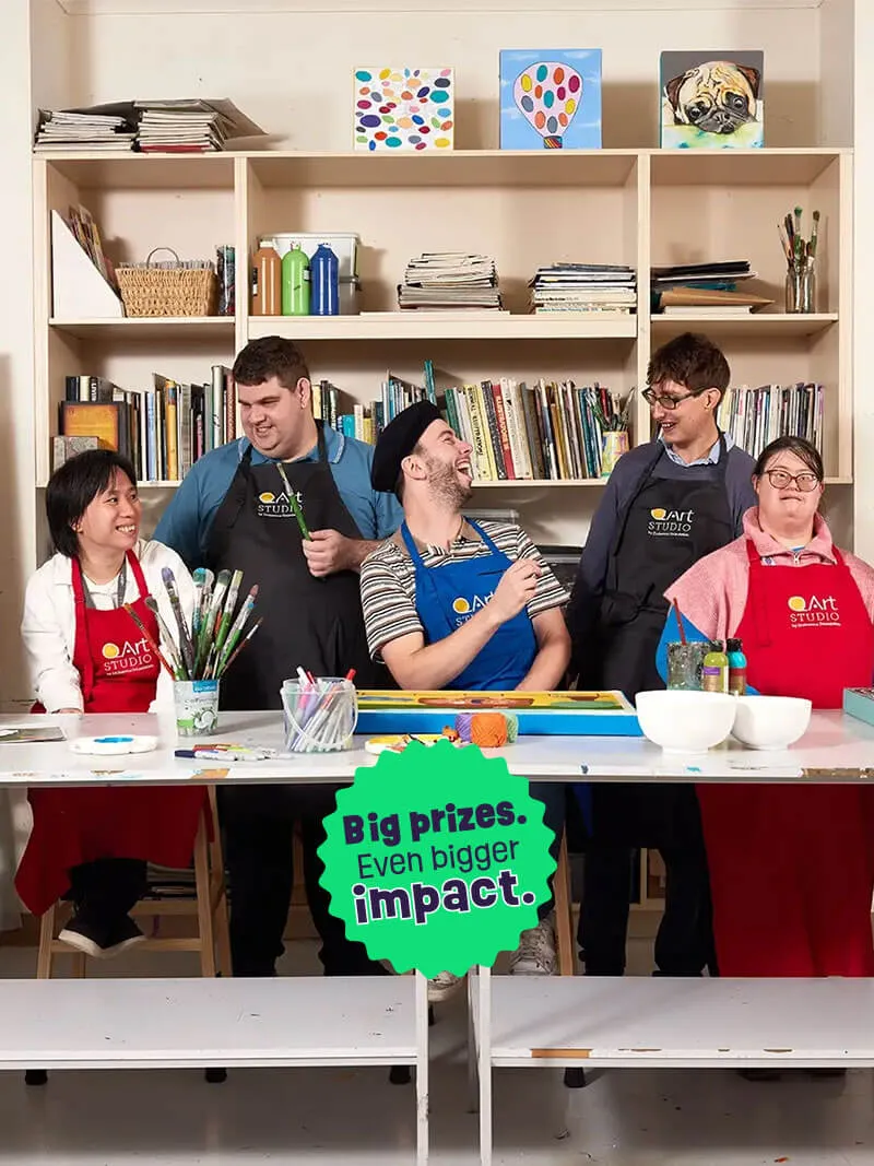 Group of five diverse adults smiling and working together in an art studio with paintbrushes, art supplies, and colorful paintings on shelves behind them.