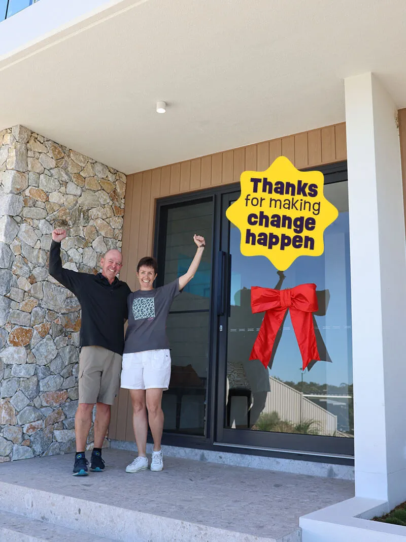 Smiling man and woman raising their fists in celebration outside a modern house entrance with a glass door decorated with a red bow and a yellow sign reading 'Thanks for making change happen.'