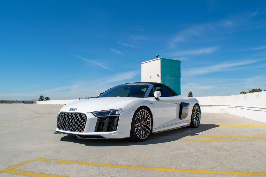 White Audi R8 V10 Spyder, front three quarter with the roof up on an open rooftop, the $795 a day car behind what it costs to rent an Audi