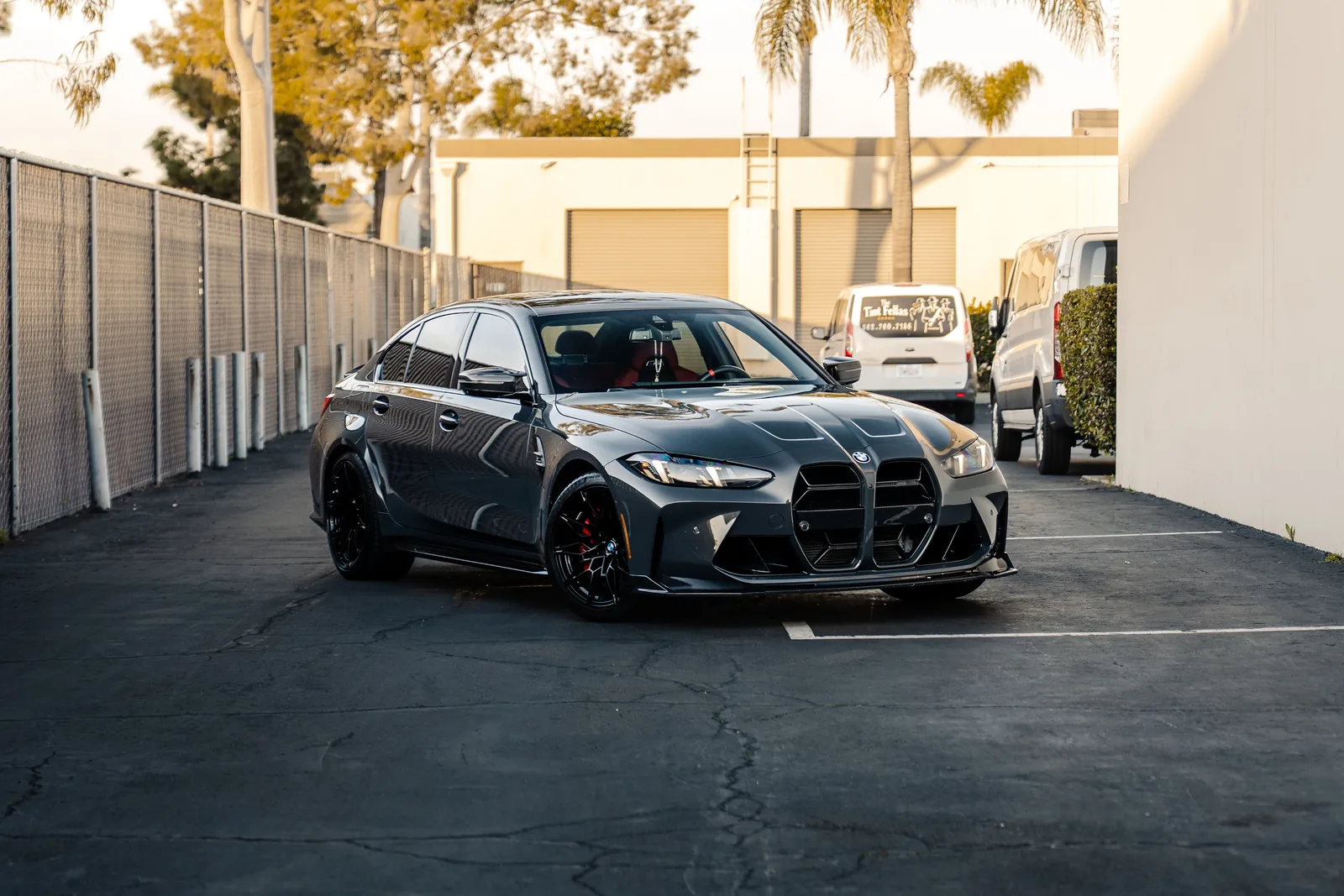 Dark grey BMW M3 Competition xDrive front three-quarter in an industrial yard beside a chain-link fence