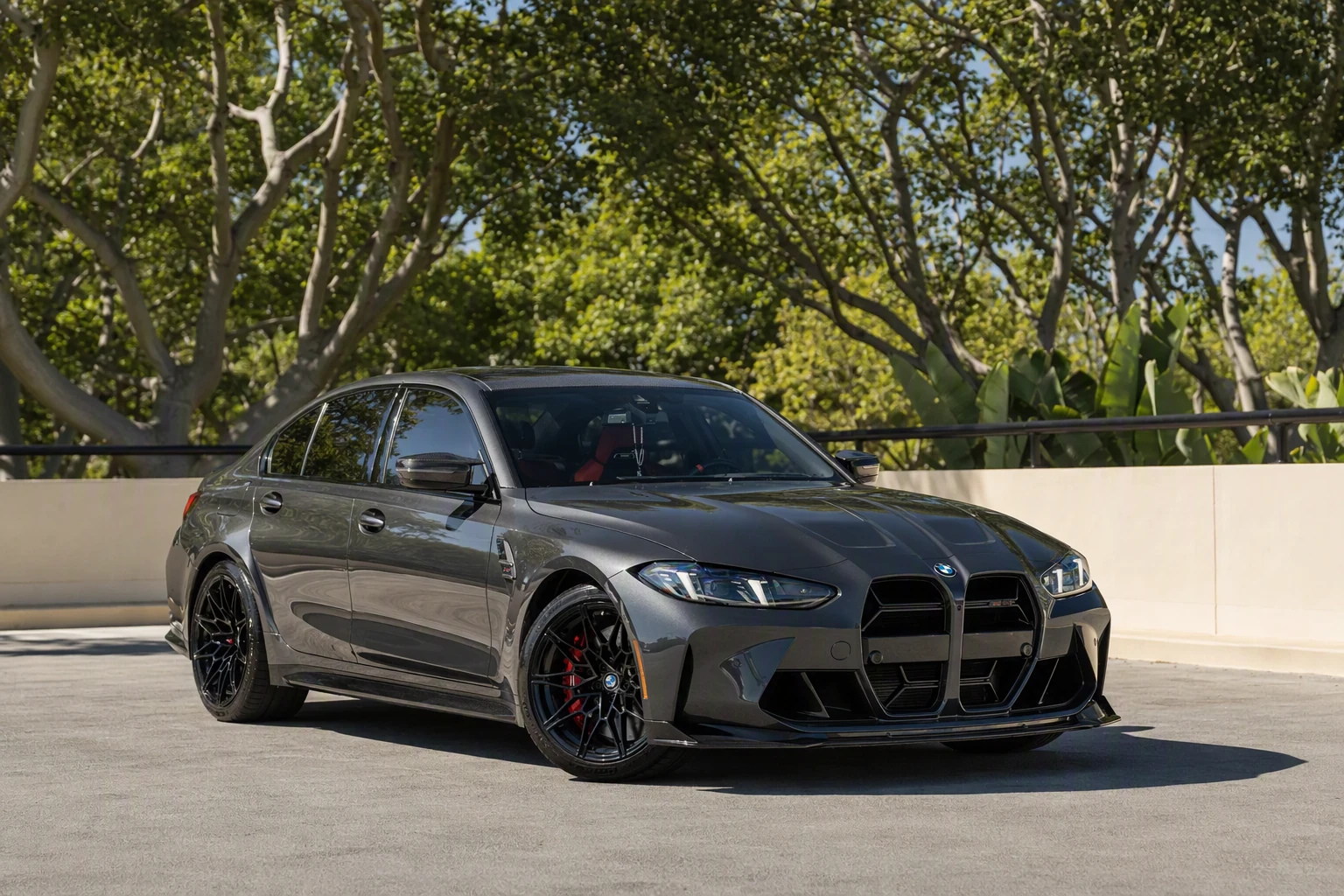 Dark grey M3 Competition xDrive parked in a tree-lined lot, black wheels and red brake calipers in the sun