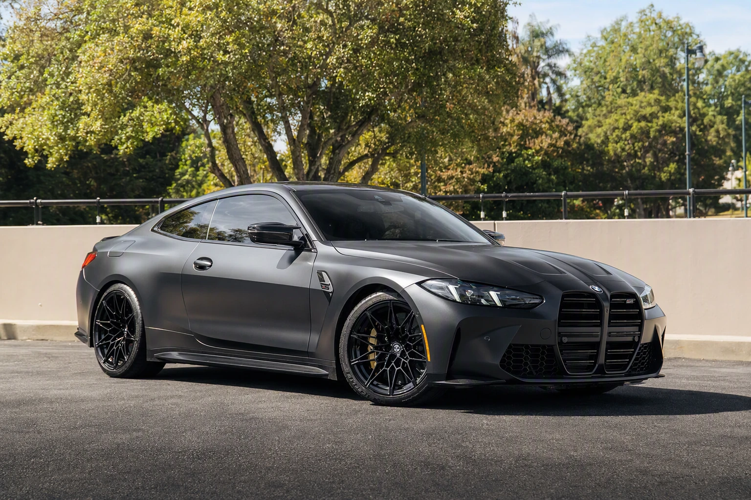 Satin black BMW M4 Competition xDrive front three-quarter under trees, yellow calipers inside black wheels