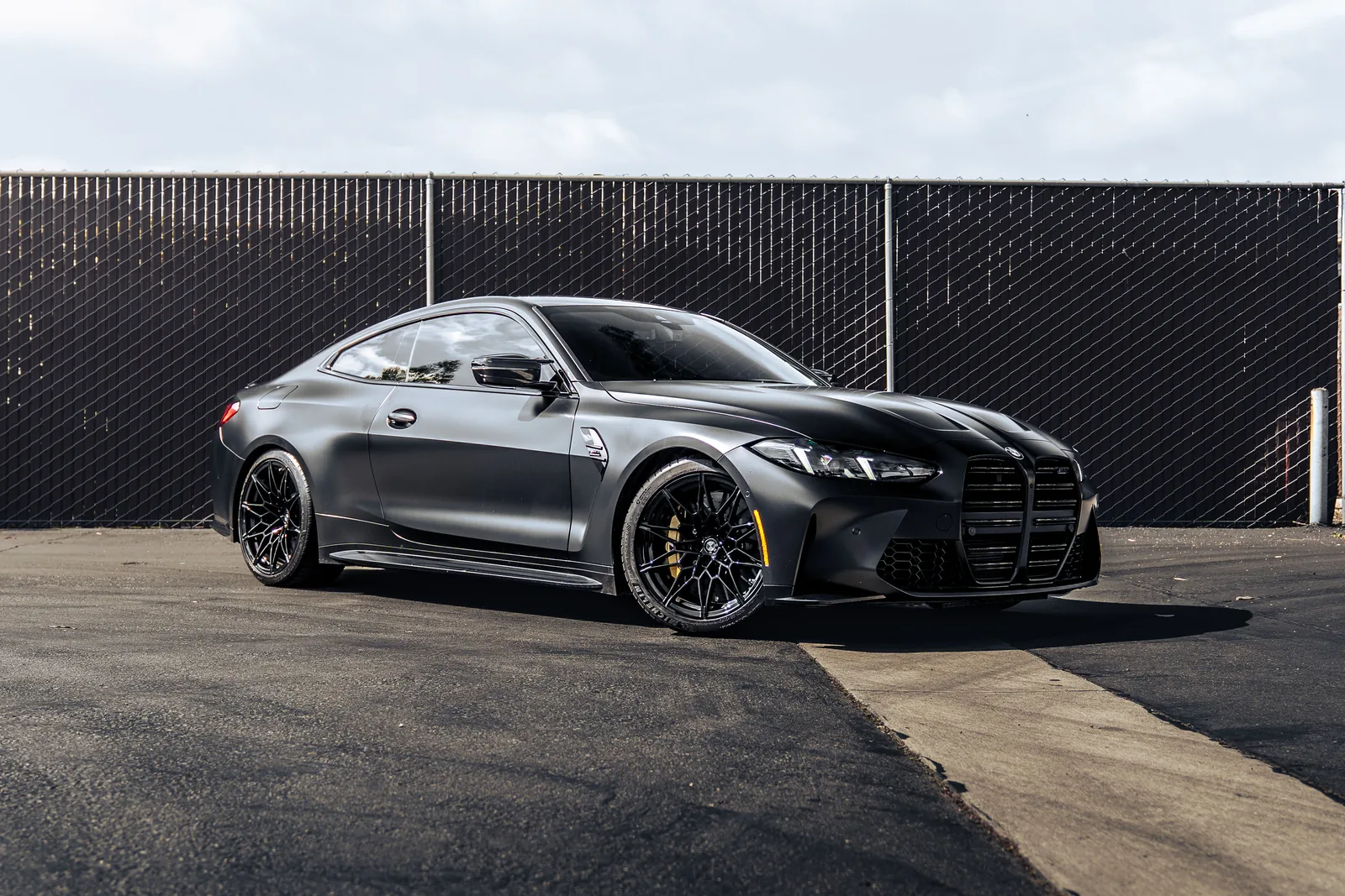 Front three-quarter of the matte black BMW M4 Competition parked by a fence on a sunny day