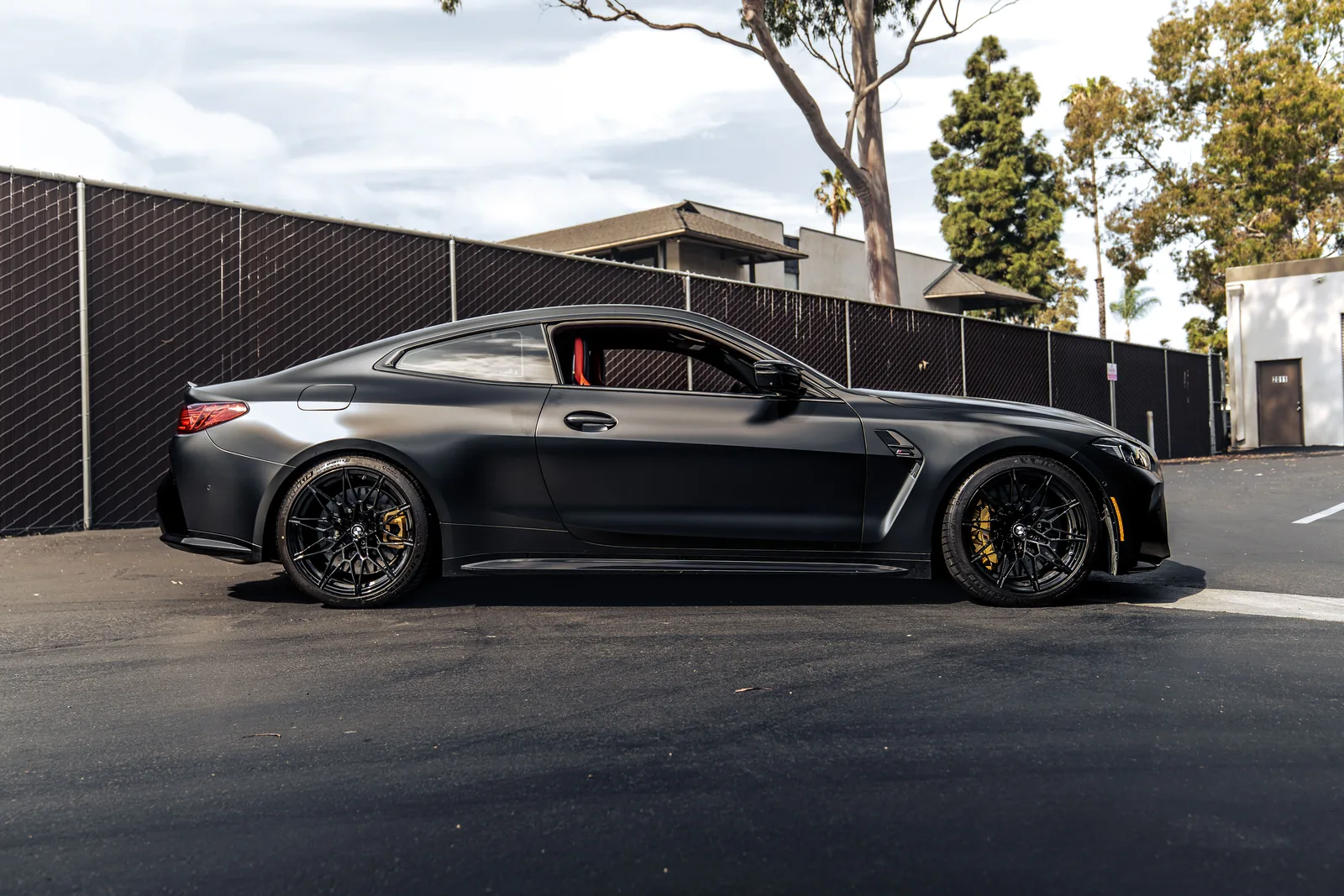 Satin black M4 Competition xDrive from the rear three-quarter beside a black fence in a parking lot