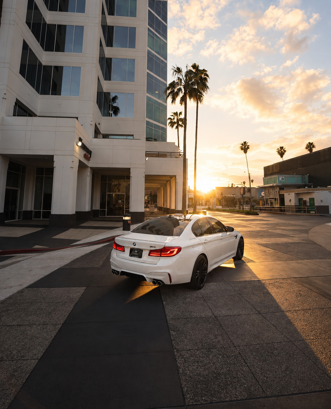 Rear view of the white M5 Competition in a plaza at golden hour, quad exhaust tips, tail lights on