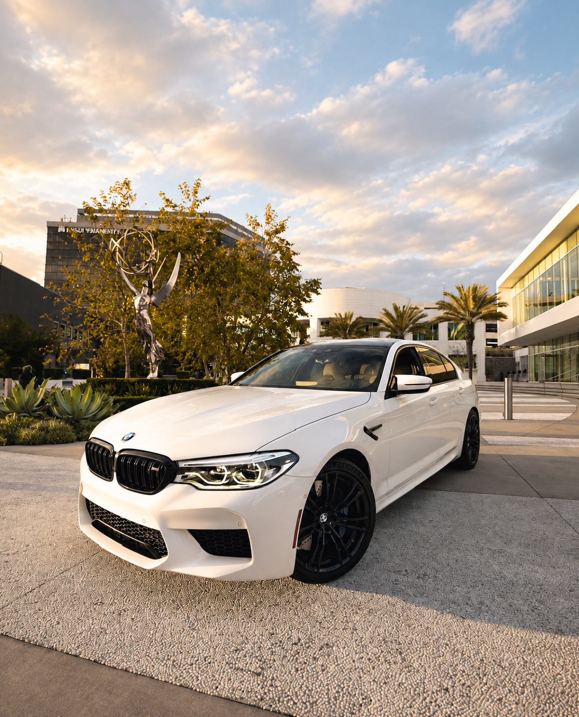 White M5 Competition front three-quarter on a plaza beside trees and a modern building