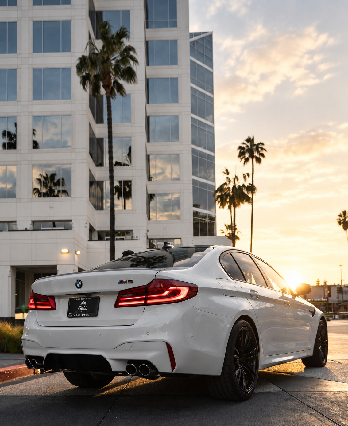 Rear three-quarter of the white M5 Competition at sunset with a glass tower and palms behind it