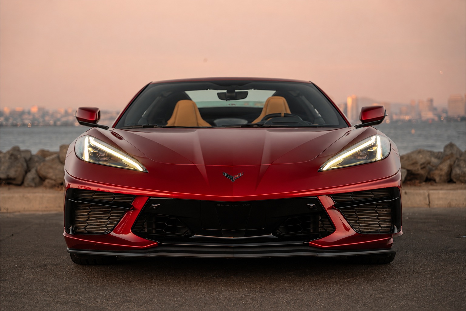 Head-on view of the red Corvette C8 Stingray Convertible at dusk, headlights on, tan seats visible over the hood