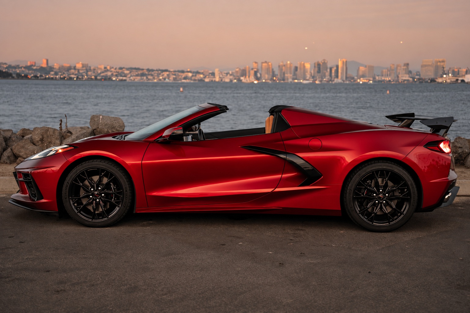 Side profile of the red Corvette Stingray Convertible, top down, high rear wing, San Diego skyline behind