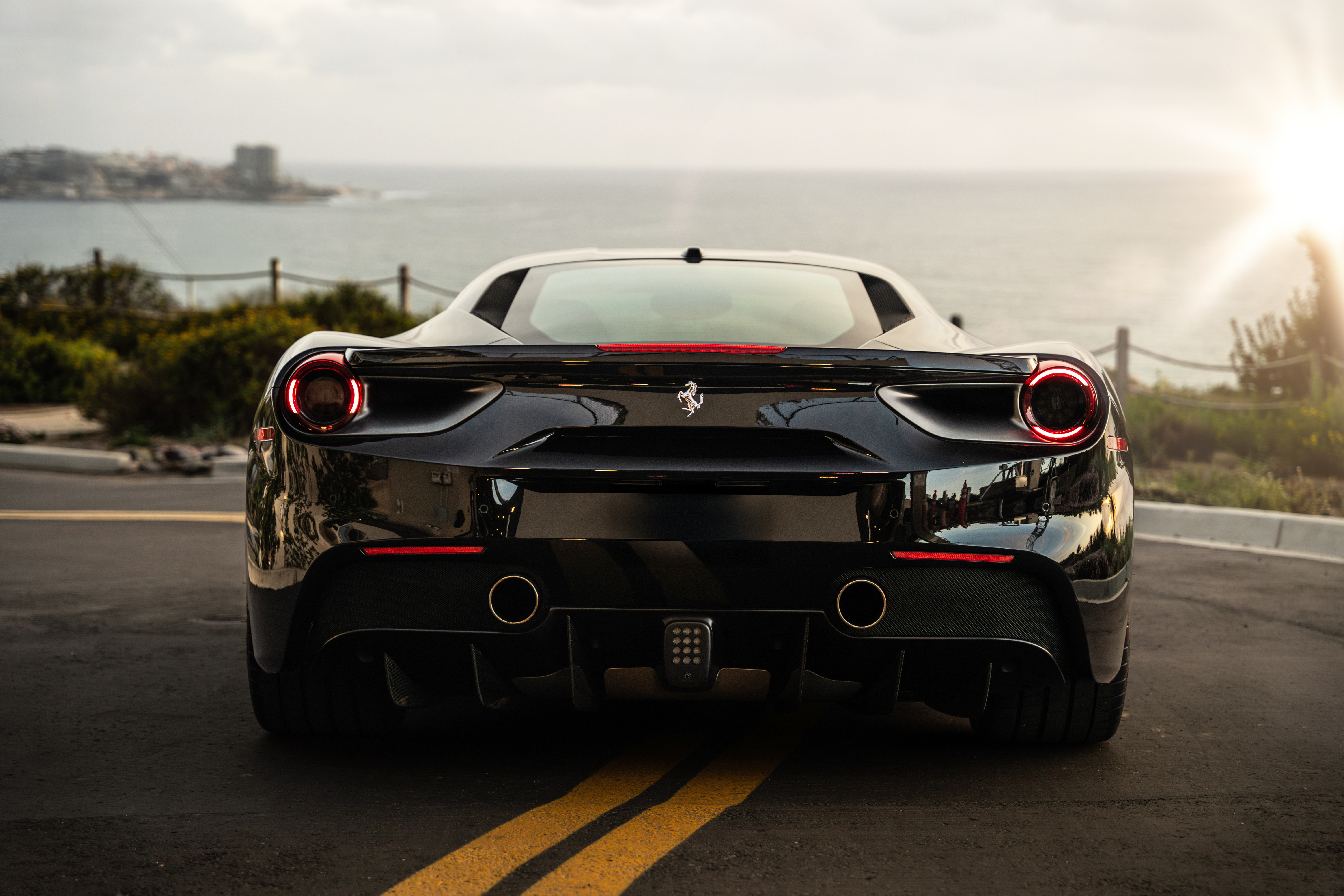 Rear view of the black Ferrari 488 GTB, round taillights and quad exhaust, coast road and ocean behind