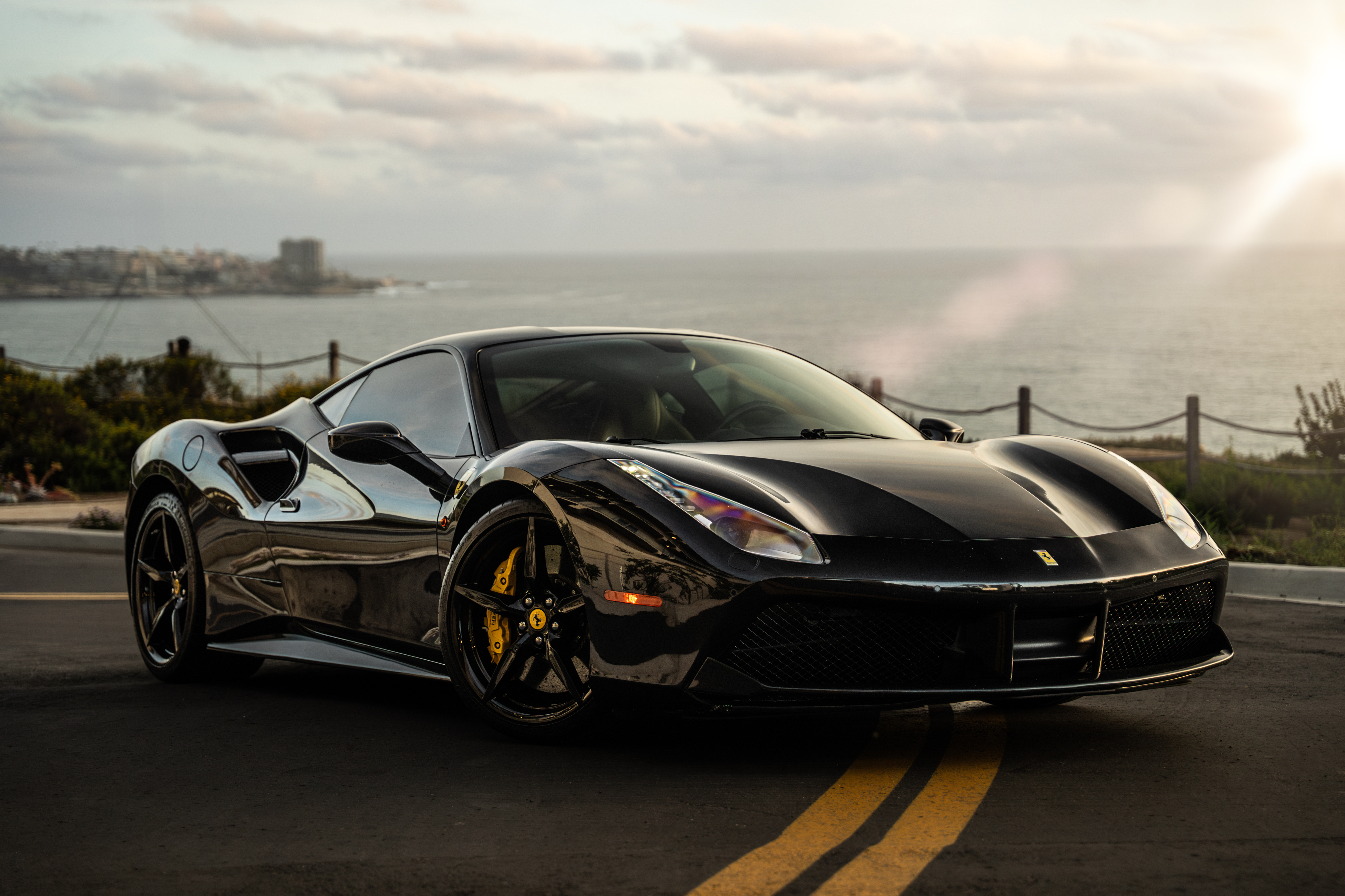 Black Ferrari 488 GTB front three-quarter on a cliff road above the ocean on the San Diego coast at sunset