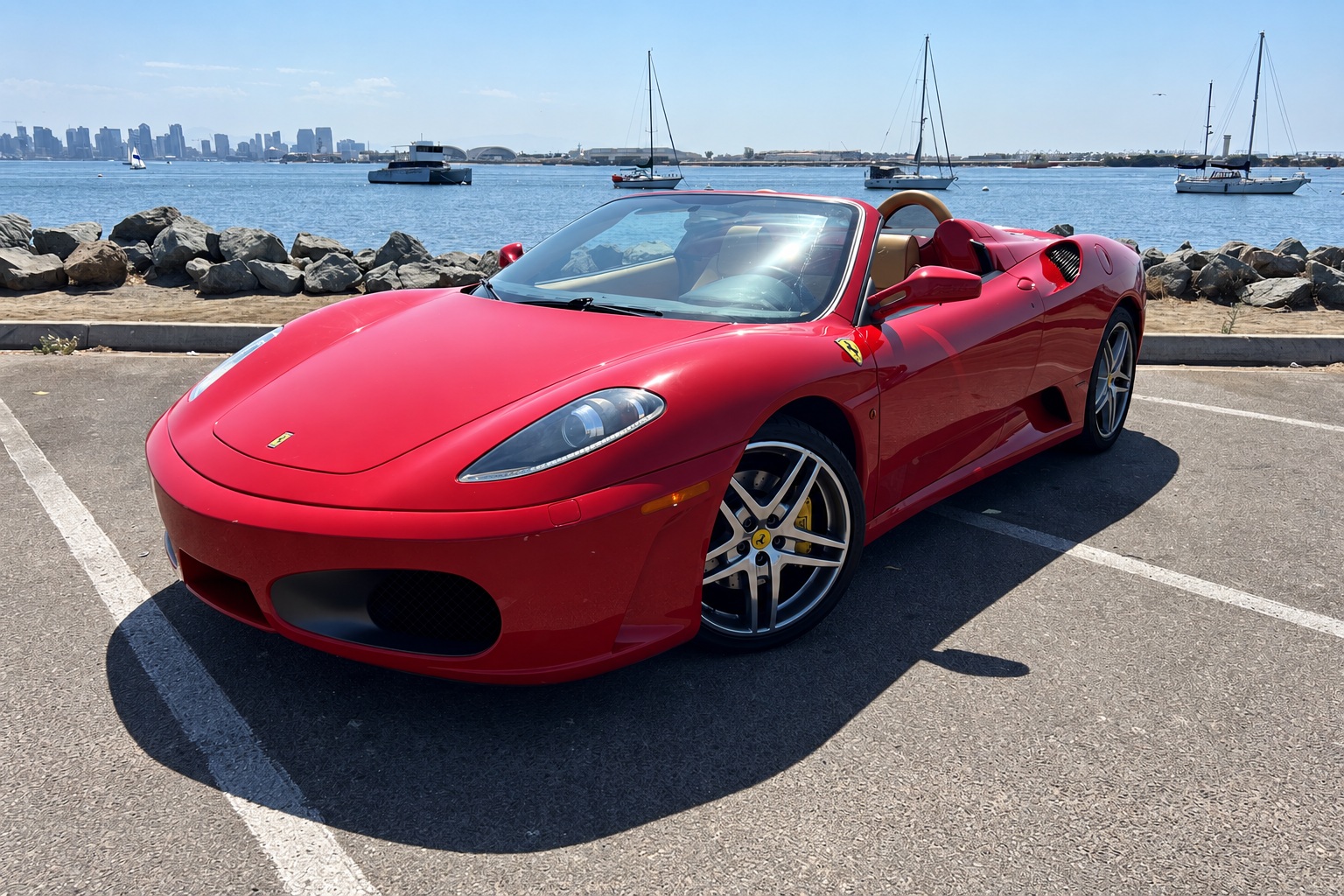 Front three-quarter of the red Ferrari F430 Spider on a waterfront lot, sailboats and a city skyline behind