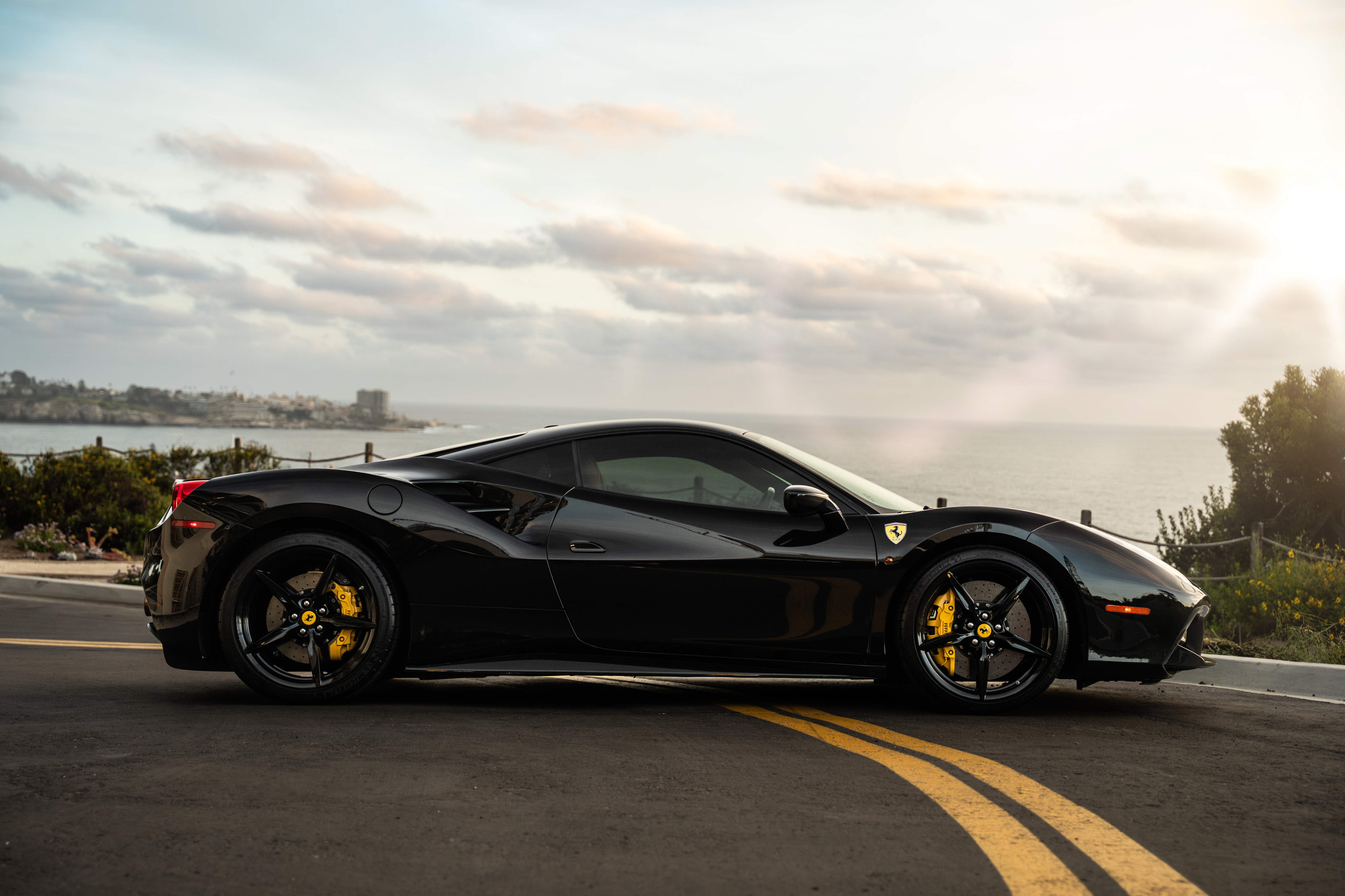 Side profile of the black Ferrari 488 GTB above the Pacific, yellow calipers behind the black 5-spoke wheels