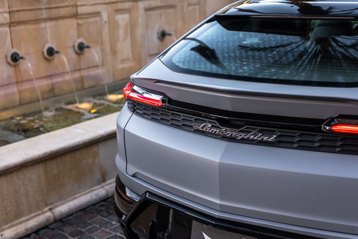 Rear of the grey Urus SE with the roof spoiler and full-width tail light beside a stone fountain